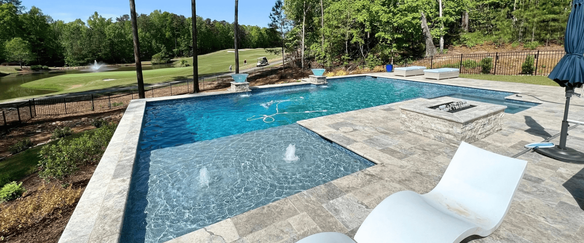 High elevated view of a custom geometric gunite pool with a shallow tanning ledge, water bubblers, in-pool lounger, raised spa, fire pit, and travertine paver deck overlooking a golf course.