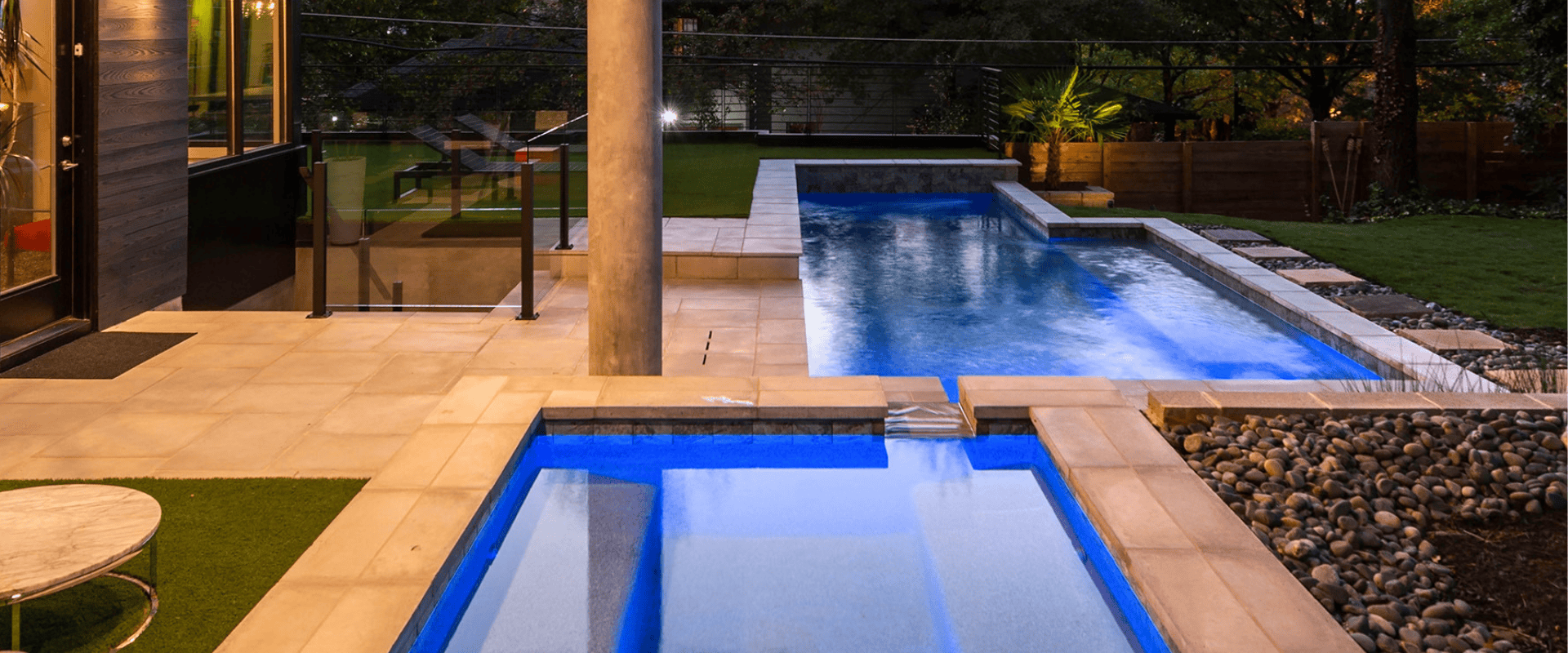 rcsoctblog2 Nighttime view of a modern geometric pool and integrated spa illuminated with blue LED lighting, featuring modern paver decking and warm home exterior illumination.